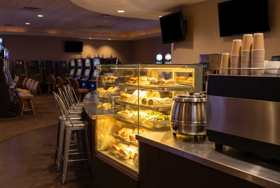 Generated illustration: a lit casino deli counter at night with sandwiches and wraps in a glass case, a soup kettle and stacked coffee cups; not a photograph of the casino.