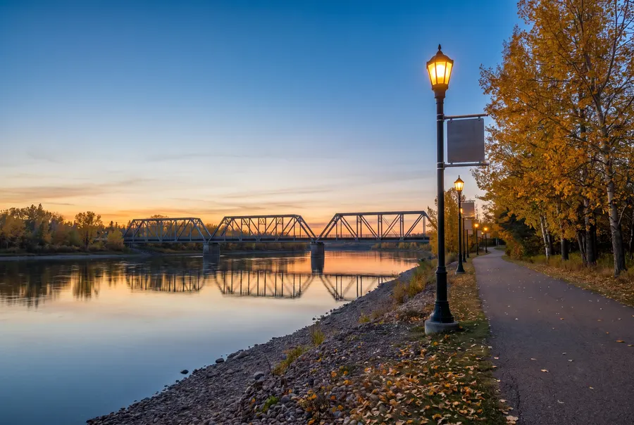 Generated illustration: a wide prairie river at dusk with a long steel truss bridge, a lamplit riverside trail and golden autumn poplars; not a specific place.