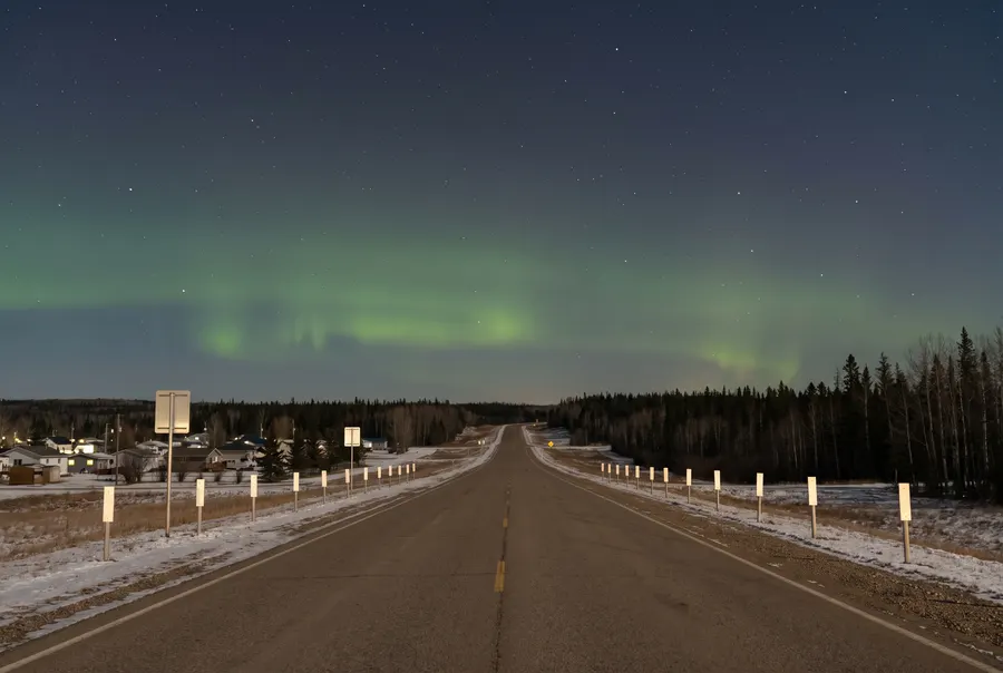 Generated illustration: a straight two-lane highway leaving a small prairie city at night towards dark forest, with green northern lights low in the sky; not a specific road.